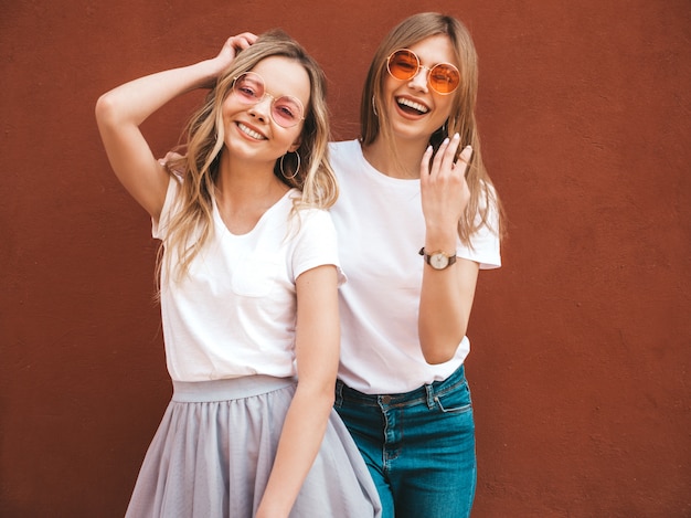 Dwie Młode Piękne Blond Uśmiechnięte Dziewczyny Hipster W Modne Letnie Białe Ubrania T-shirt. Kobiety Pozuje W Ulicznej Pobliskiej Czerwieni ścianie. Pozytywne Modele Zabawy W Okularach Przeciwsłonecznych Darmowe Zdjęcia