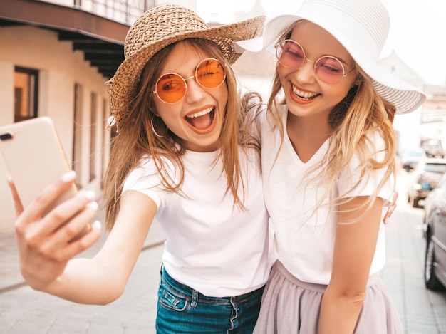 Dwie Młode Uśmiechnięte Hipster Blond Kobiety W Letniej Białej Koszulce. Dziewczyny, Robiąc Selfie Autoportret Zdjęcia Na Smartfonie. Modele Pozowanie Na Tle Ulicy. Kobieta Pokazuje Pozytywne Emocje Darmowe Zdjęcia