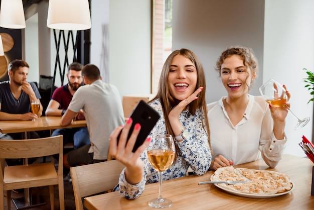 Dwie pozytywne dziewczyny uśmiecha się i robi selfie z winem w restauracji. Premium Zdjęcia