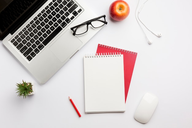 Eyeglasses na laptopie, jabłku, słuchawkach, barwionym ołówku, ślimakowatym notepad i myszy na białym biurku Darmowe Zdjęcia