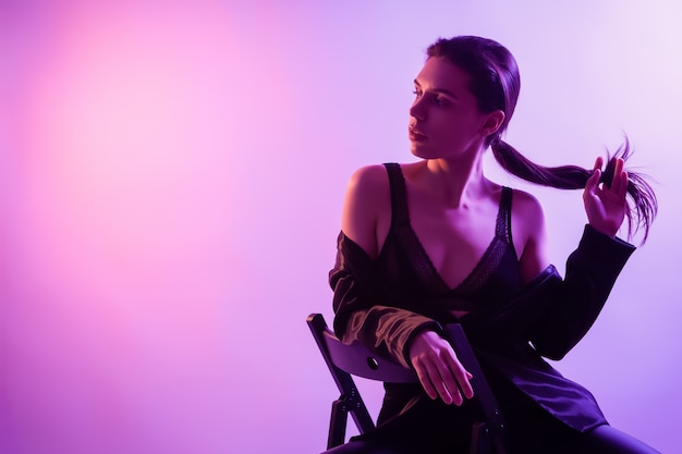 Filmowa Noc Portret Kobiety W Neonie. Piękna Młoda Kobieta W Stylowe Ubrania Pozowanie Na Krześle W Kolorowe światła. Premium Zdjęcia