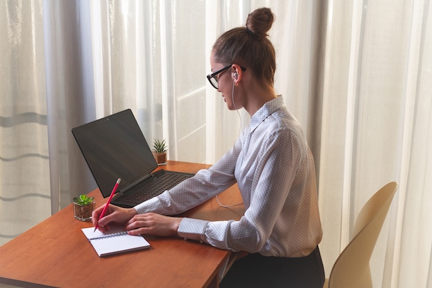 Młoda kobieta w słuchawkach angażowała się w kursy online zdalnie, używając laptopa i zapisując informacje w notatniku Premium Zdjęcia