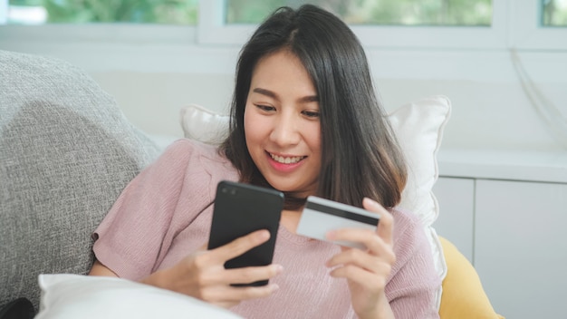 Młoda uśmiechnięta azjatycka kobieta używa smartphone kupuje online zakupy kredytową kartą podczas gdy kłamający na kanapie gdy relaksuje w żywym pokoju w domu. styl życia kobiety pochodzenia łacińskiego i latynoskiego w domu koncepcja Darmowe Zdjęcia