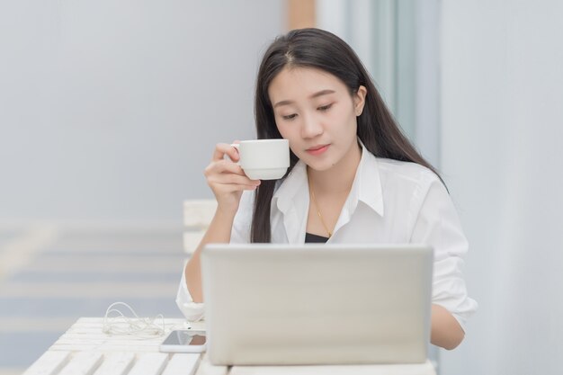Portret uroczej azjatyckiej modelki używa laptopa do komunikacji online; szczęśliwej biznesowej kobiety napoju kawowy obsiadanie przy białym biurkiem. Premium Zdjęcia