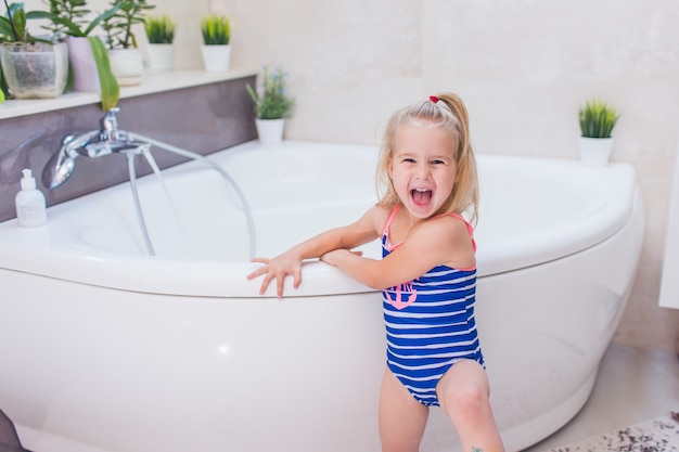 Szczęśliwa Mała Dziewczynka W Whetu Błękitnym Swimsuit Zostaje Blisko Wanny W łazience I Krzyczy Z Uśmiechem. Premium Zdjęcia