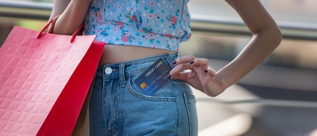 Szczęśliwa młoda kobieta trzyma kartę kredytową z kieszeni dżinsów z torby na zakupy, wydaje pieniądze z cieszyć się na zakupy. Premium Zdjęcia