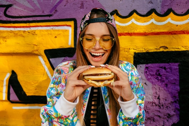 Szczęśliwa piękna młoda kobieta, ono uśmiecha się szeroko, trzyma smakowitego hamburger w dwa rękach. Darmowe Zdjęcia