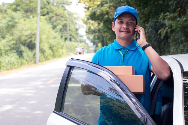 Alegre repartidor asiático sosteniendo una caja y sonriendo mientras está de pie en la carretera Foto Premium