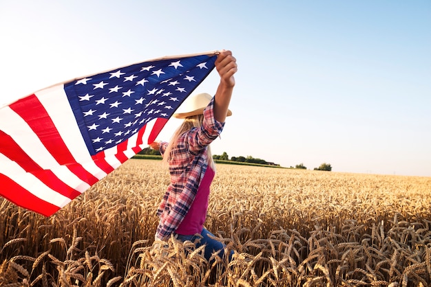 Atractiva mujer rubia caminando por el campo de trigo y sosteniendo la bandera ondeante de estados unidos Foto gratis