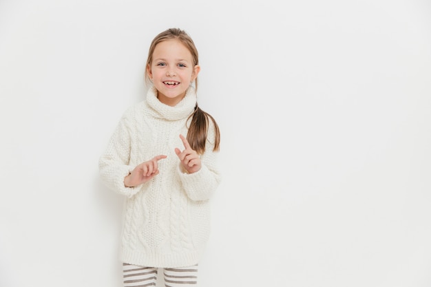 Atractiva niña pequeña en suéter de invierno cálido, escucha una historia positiva de un amigo, se levanta contra blanco Foto Premium