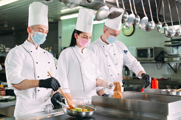 Los Chefs Con Mascaras Protectoras Y Guantes Preparan La Comida En La Cocina De Un Restaurante U Hotel Foto Premium