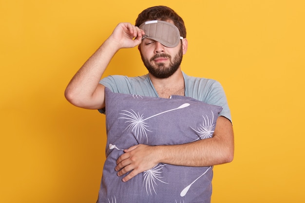 Closeup retrato de hombre soñoliento con los ojos vendados en los ojos, sosteniendo la almohada en las manos, abre la máscara para dormir, manteniendo los ojos cerrados Foto gratis