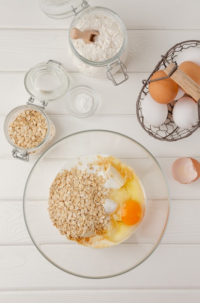 Cocinar panqueques de avena con plátanos. huevos, suero de leche, azúcar, vainilla, avena en un recipiente sobre un fondo de madera blanca. receta paso a paso. Foto Premium