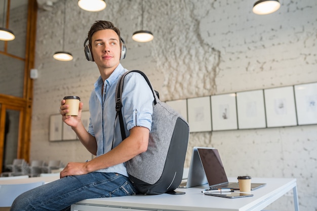 Cool apuesto joven sentado en la mesa en auriculares con mochila en la oficina de coworking tomando café, | Foto Gratis