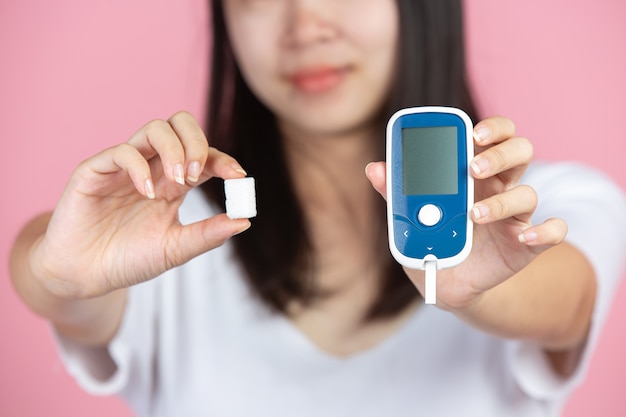 Día mundial de la diabetes; mujer sosteniendo medidor de glucosa y terrones de azúcar en la pared rosa Foto gratis