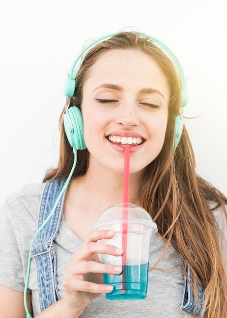 Escuchar música de mujer en los auriculares beber jugo con paja contra el fondo blanco Foto gratis