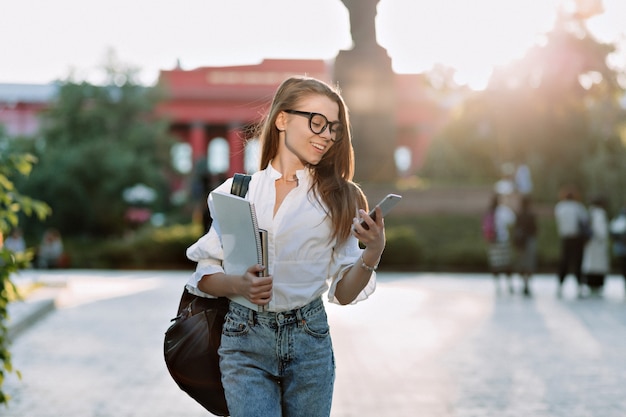 Estudiante morena sonriente con gafas y camisa caminando al aire libre con una computadora portátil y una mochila y mirando en el teléfono inteligente en la luz del sol Foto gratis