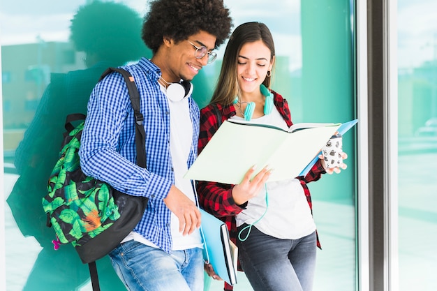 Estudiantes adolescentes con libros y sus bolsas de pie contra el vidrio mirando el libro Foto gratis