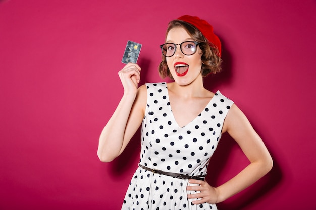 Feliz mujer de jengibre en vestido y anteojos con tarjeta de crédito mientras mira a la cámara sobre rosa Foto gratis