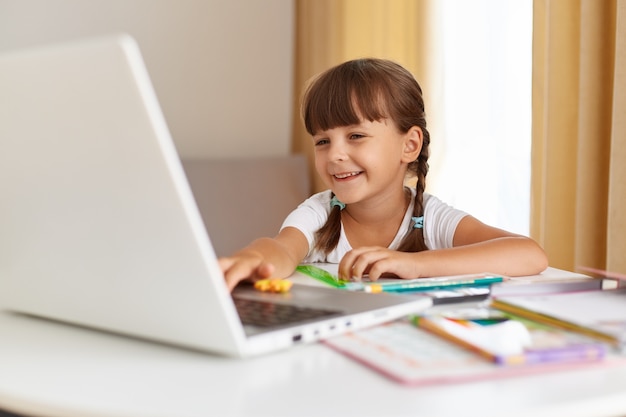 Filmación en interiores de un alumno de pelo oscuro positivo feliz posando en casa, mirando la computadora portátil con una sonrisa encantadora, educación a distancia, con lecciones en línea. Foto gratis