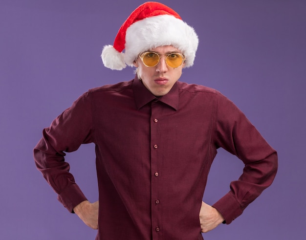 Fruncir el ceño joven rubio con gorro de papá noel y gafas manteniendo