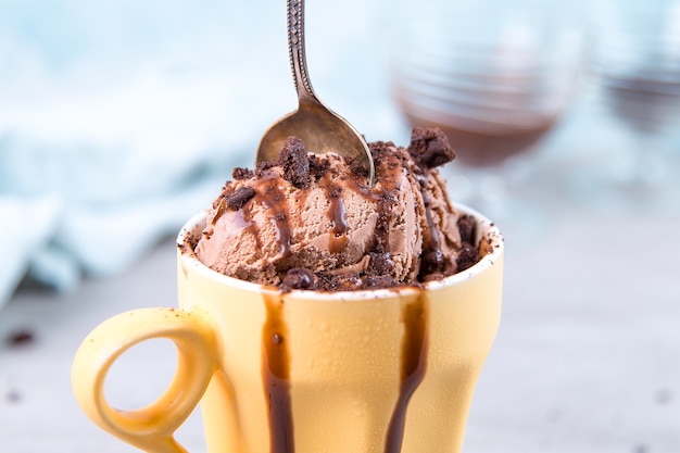 Helado de chocolate marrón en una taza y cuchara de color amarillo normal, sobre azul, con cobertura y migas. Foto Premium 