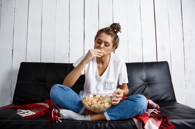 Hermosa mujer doméstica comiendo patatas fritas, viendo la televisión, sentado en el sofá. Foto gratis