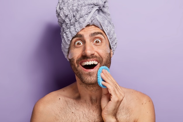 El Hombre Alegre Y Sorprendido Quiere Tener Una Piel Suave Se Limpia La Cara Con Una Esponja Cosmetica Se Aplica Parches De Silicona Debajo De Los Ojos Foto Gratis