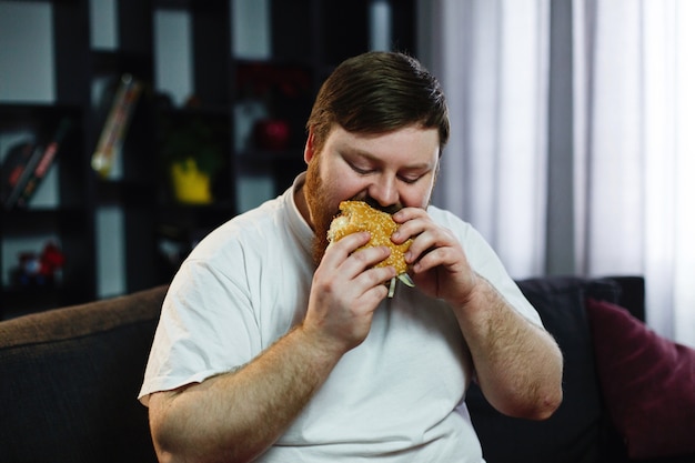El hombre gordo sonriente come la hamburguesa que se sienta antes de un televisor Foto gratis