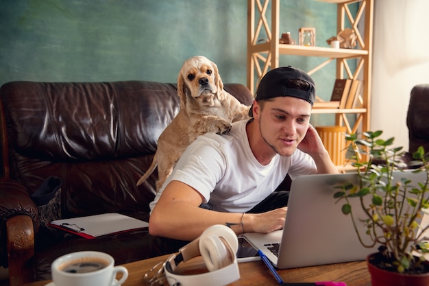 Hombre guapo joven sentado en el sofá marrón y trabajando con su lindo perro Foto gratis
