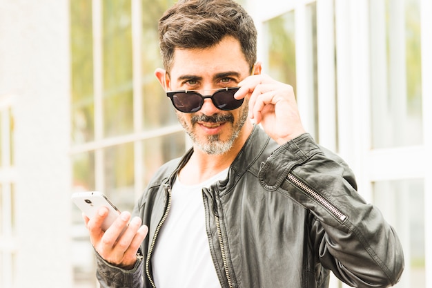 Hombre hermoso que mira a escondidas a través de las gafas de sol que sostienen el teléfono móvil Foto gratis