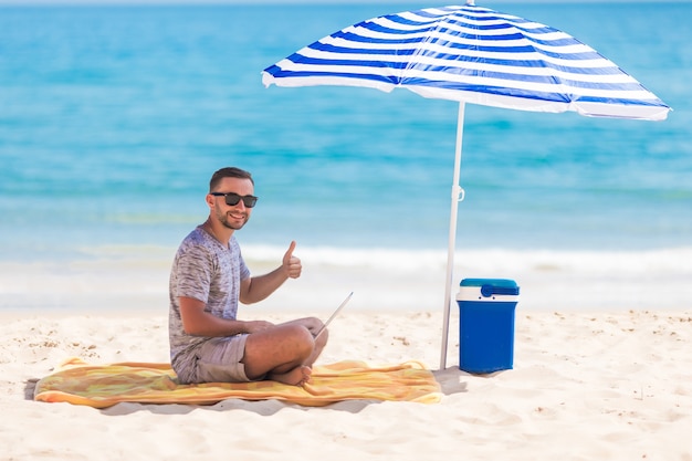 Hombre joven feliz en la playa bajo la sombrilla cerca del océano trabajando en su computadora portátil y mostrando los pulgares para arriba Foto gratis
