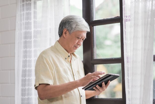 Hombre Mayor Asiatico Que Usa La Tableta Que Comprueba Medios Sociales Cerca De Ventana En Sala