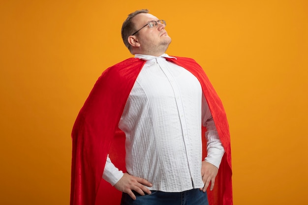 Hombre de superhéroe adulto seguro en capa roja con gafas manteniendo las manos en la cintura mirando al lado aislado en la pared naranja Foto gratis