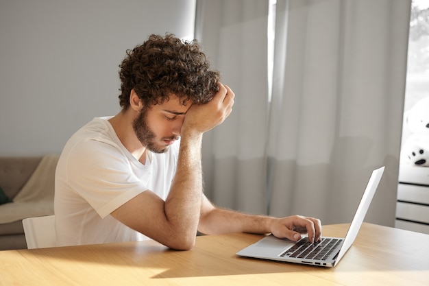 Imagen de un traductor joven cansado con barba espesa y cabello ondulado tocando la cabeza y cerrando los ojos, sintiéndose absolutamente exhausto, escribiendo en el teclado de la computadora portátil, traduciendo documentación técnica Foto gratis