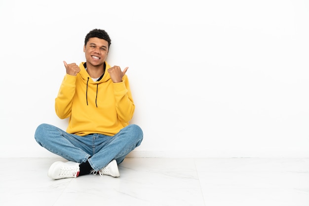 Joven Afroamericano Sentado En El Suelo Aislado Sobre Fondo Blanco Con Gesto De Pulgar Hacia 1884