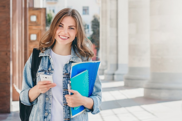 Joven estudiante sonriente contra la universidad. linda chica estudiante  tiene carpetas, cuadernos y teléfono móvil en las manos. aprendizaje,  educacion | Foto Premium