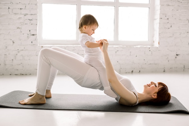 Joven Madre Deportiva Hace Ejercicios Fisicos De Yoga O Pilates Puente Trasero Junto Con Su Bebe Contra El Fondo De Grandes Ventanales Fitness Feliz Maternidad Foto Premium