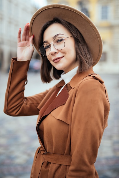 Joven Mujer Bonita En Gafas Modernas Y Sombrero De Moda Y Abrigo Marron Esta Posando En El Centro De La Ciudad Foto Gratis