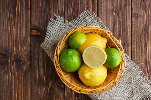 Limas en una canasta de mimbre con limones planos sobre madera y un saco Foto gratis