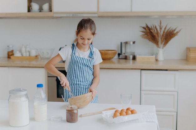 Linda Nina Pequena Usa Un Delantal A Rayas Bate Los Ingredientes En Un Tazon Prepara La Masa Ensena A Cocinar Foto Premium