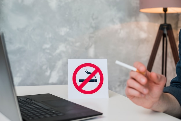 Mano del hombre que sostiene el cigarrillo en la computadora portátil frontal y no fumar signo Foto gratis