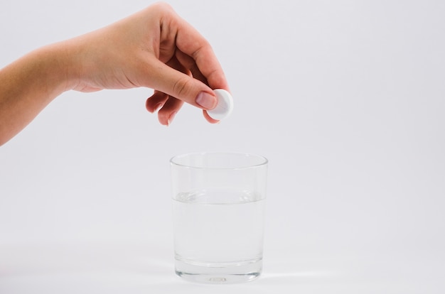 Mano de la mujer sosteniendo la píldora blanca sobre el vaso de agua contra el fondo gris Foto gratis