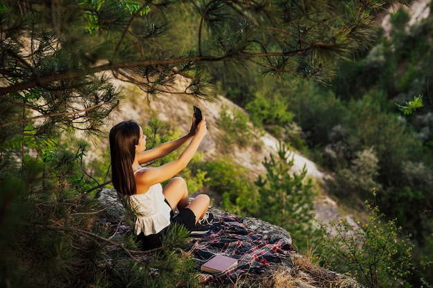 Mujer atractiva joven sosteniendo un teléfono inteligente y tomando un selfie en la roca, sobre el valle con un bosque de pinos perfecto. Foto Premium 