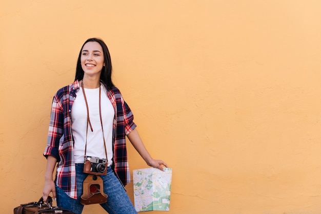 Mujer bastante joven feliz que sostiene el bolso y el mapa que se colocan cerca de la pared del melocotón Foto gratis