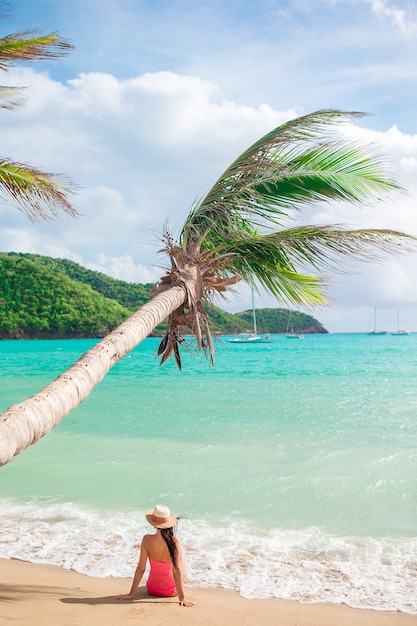 Mujer delgada joven en el sombrero del bikini y de paja que miente en la playa tropical. hermosa chica bajo la palmera en aguas poco profundas Foto Premium
