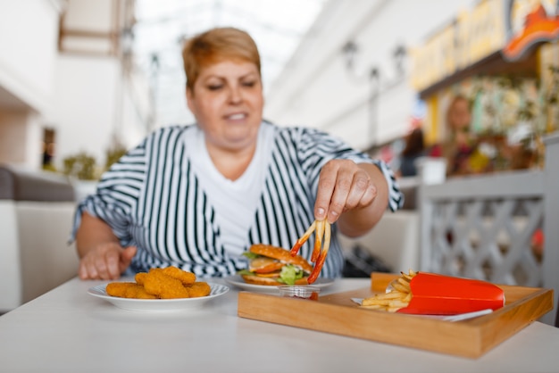 Mujer gorda comiendo papas fritas en el patio de comidas del centro comercial. persona de sexo femenino con sobrepeso en la mesa con comida chatarra, problema de obesidad Foto Premium 