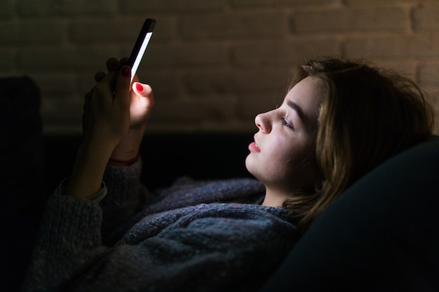 Mujer hermosa joven que usa el teléfono inteligente por la noche en la cama - tecnología, red social, concepto de insomnio Foto gratis