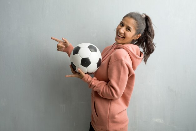 Mujer India Deportiva Joven Contra Una Pared Que Senala Al Lado Sonriendo Sorprendida Presentando Algo Natural Y Casual Sosteniendo Una Pelota De Futbol Para Jugar Un Juego Foto Premium