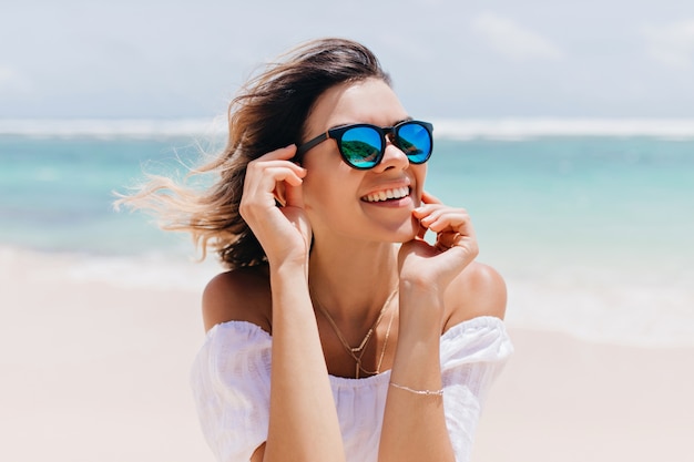 Mujer maravillosa en traje blanco y gafas brillantes posando con expresión de cara feliz en un caluroso día de verano. agradable mujer caucásica de pie cerca del océano en el cielo Foto gratis